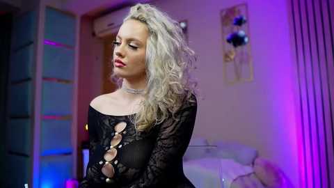 Video of a pale, platinum blonde woman with wavy hair, wearing a black lace top with cutouts, standing in a dimly lit room with neon lights.