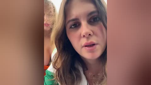 Video of a young woman with long, wavy brown hair, light skin, and a distressed expression, wearing a green and white top. Background is blurred, showing a beige and orange surface.