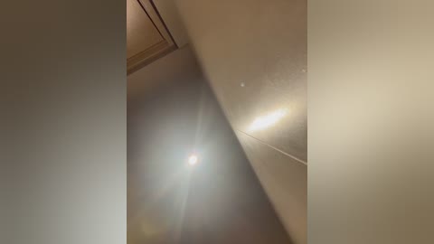 Media: Video of a beige wall with a glossy finish, featuring a lens flare from a light source above. The image is slightly out of focus, emphasizing the reflective quality of the wall and the bright spot.