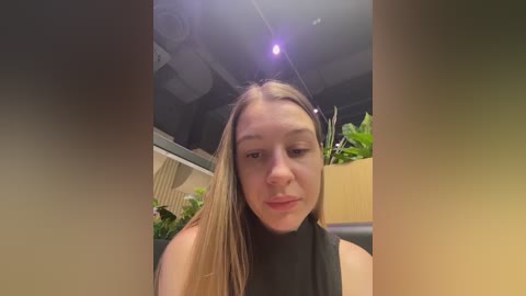 Media: Video of a young Caucasian woman with long blonde hair, wearing a black turtleneck, standing indoors with plants in the background.