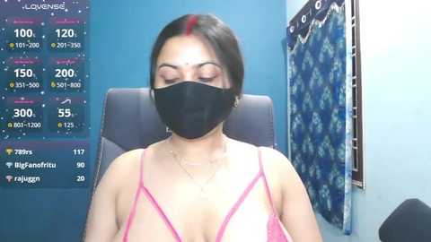 Video of a South Asian woman with fair skin, wearing a black face mask, pink spaghetti-strap top, and gold necklace, sitting in a chair, background showing cryptocurrency exchange screen and blue floral curtains.