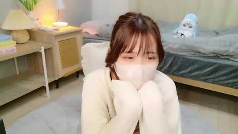 Media: Video of a young Asian woman in a white mask, sitting on a light carpet, wearing a beige sweater, in a modern bedroom with a gray bed and stuffed animal.