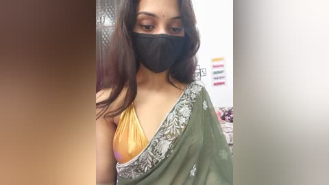 Video of a South Asian woman with medium-brown skin, wearing a yellow blouse and a green sari with intricate silver embroidery, a black face mask, and long dark hair, indoors with a blurred background.