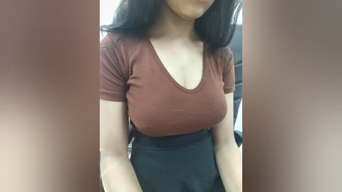 Media: Video of a young woman with medium brown skin, wearing a tight, ribbed brown V-neck top that accentuates her medium-sized breasts and high-waisted black skirt. She has long, straight black hair. Background is blurred, suggesting an indoor setting.