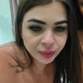 Video of a young woman with fair skin, long dark hair, and full lips, wearing dark eyeliner, pouting. She has a tattoo on her shoulder and is topless. Background is blurred.