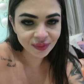 Video of a young woman with long dark hair, wearing dark eyeliner, and red lipstick, smiling. She has a tattoo on her left shoulder. Background shows a blurred bed with white sheets.