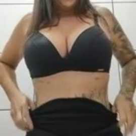 Video of a curvy, tattooed woman with medium-length dark hair, wearing a black bra and black pants, posing in front of a white tiled wall.