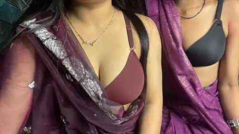 Video of two South Asian women with medium to dark skin tones wearing purple sarees. One has a burgundy bra visible, the other a black one, both with medium-sized breasts. They are indoors, likely in a private setting.