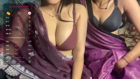 Video of two women, one in a purple satin sari with a black bra, the other in a black bra, both with large breasts, sitting on a bed with colorful bedding.
