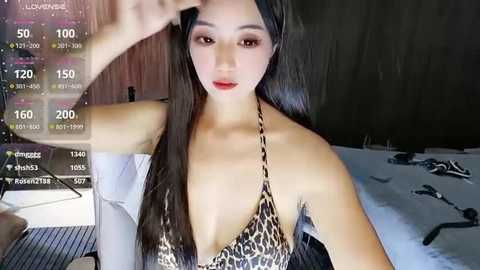 Video of an East Asian woman with long black hair, wearing a leopard-print halter top, posing in a bedroom with dim lighting.
