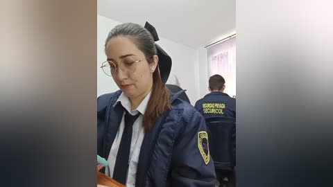 Media: Video of a young woman with glasses, fair skin, and dark hair tied back, wearing a navy-blue police uniform with a black tie, standing in a room with a colleague.