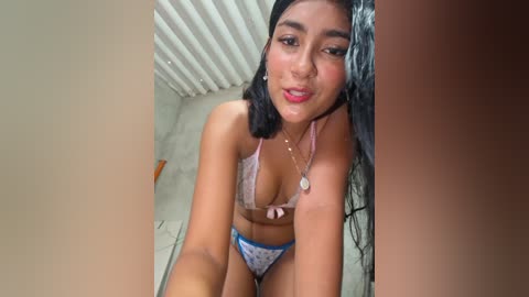 A video of a young Latina woman with medium brown skin, long black hair, wearing a light pink bra and white polka dot panties, kneeling in a shower with water running, smiling.
