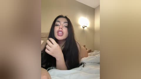 Video of a young woman with long black hair, wearing a black top, lying on a bed with beige walls and a wall-mounted lamp in the background.