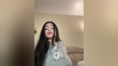 A video of a young woman with long, dark hair, wearing a light gray hoodie with a graphic design, leaning against a beige wall. She has a slight smile and is in a casual, indoor setting.