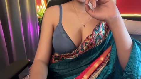 A video of a woman in a teal sari with a blue bra, resting her chin on her hand, in a dimly lit room with red and purple curtains.
