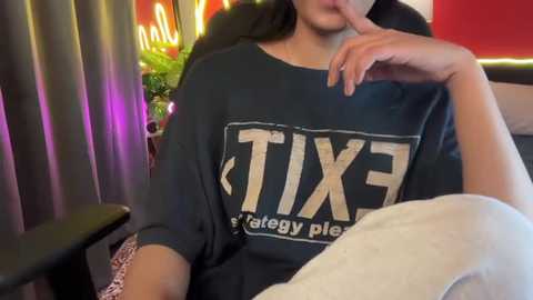 Video of a woman with long dark hair, wearing a large, dark gray \"TIX\" t-shirt, sitting on a black chair. She has her finger to her lips, in a casual, indoor setting with colorful lights and a green plant in the background.