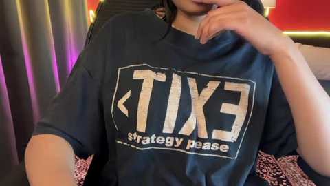 Video of a woman with light brown skin, wearing a black T-shirt with \"TIX\" and an arrow graphic, resting her chin on her hand, in a dimly lit room with red and purple lights.