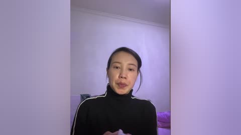 A video of an Asian woman with straight black hair, wearing a black tracksuit with white stripes, standing in a minimalist room with white walls and a purple bed.