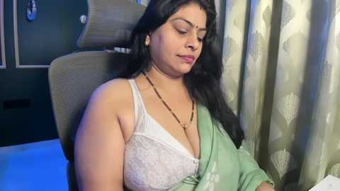 A video of a South Asian woman with long black hair, wearing a white lace bra and green shawl, sitting on a grey cushioned chair, with beige curtains in the background.