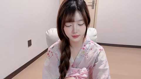 Media: Video of a young Asian woman with long brown hair in a floral kimono, seated in a minimalist room with light beige walls and a white chair.