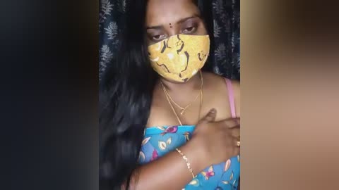 A video of a South Asian woman with medium brown skin, wearing a yellow face mask with butterfly patterns, a blue floral top, and a gold bracelet.