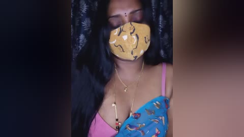 Video of a South Asian woman with long black hair, wearing a blue sari adorned with floral embroidery, a yellow mask, and a pink bra. She has a gold necklace. Background is dark and blurry.