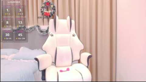A video of a plush white gaming chair with a pink pillow on the seat, situated in a cozy bedroom with beige curtains and a Christmas wreath on the wall.