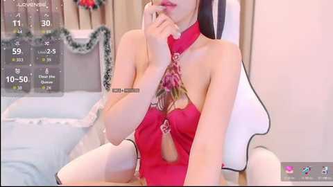 Video of an Asian woman with long black hair, wearing a revealing red halterneck dress with floral pattern, seated in a white chair. Background shows a cozy room with Christmas decorations.