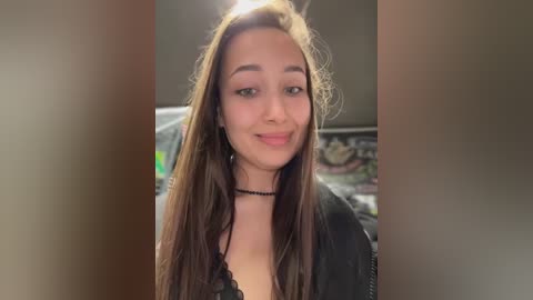 Video of a young woman with long, straight brown hair, wearing a black lace top and choker necklace. She has a light olive complexion and a slight smile. Background shows a blurred bedroom with a bed and wall decorations.