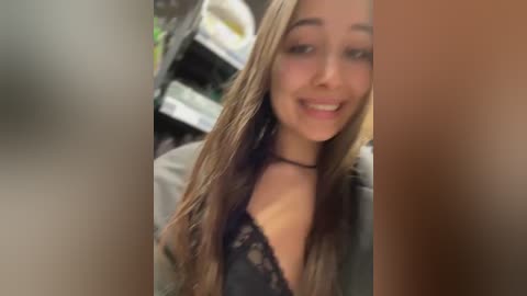 A video of a young woman with long, straight brown hair and light skin, wearing a black lace bralette and a denim jacket, smiling. The background shows a blurred kitchen with shelves and appliances.