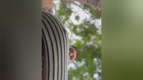 Video of a man wearing a striped shirt, seen from a low angle, with blurred green tree foliage in the background.