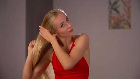Video of a slender, fair-skinned blonde woman with long hair, wearing a red sleeveless top, adjusting her hair in a room with gray walls and abstract art.