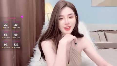 Video of a young woman with long brown hair, fair skin, and a slender figure, wearing a sleeveless beige top, sitting on a bed with a fluffy white blanket. Background shows a lamp, beige walls, and a framed picture.