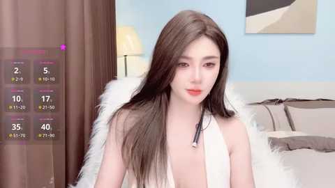 Video of a fair-skinned Asian woman with long, straight black hair, wearing a white sleeveless dress, sitting on a bed with beige pillows, in a modern bedroom with blue walls and abstract art.
