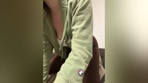 Media: A close-up video of a person wearing a green jacket with a white logo on the sleeve, sitting in a chair with a beige and brown patterned cushion. The background is plain, with soft lighting.