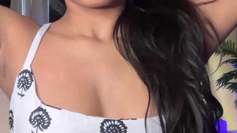Media: Video of a woman with long, dark hair, wearing a white tank top with black floral patterns. Her cleavage is visible, showing medium-sized breasts. Background includes a green plant and a blurred indoor setting.