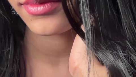 Media: Video of a close-up of a woman's face, showcasing her full lips in a glossy, pink lipstick and long, straight black hair. The background is blurred, focusing on her face.