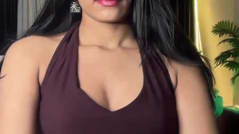 Video of a woman with medium brown skin, wearing a low-cut, dark brown halter top that highlights her ample cleavage, with long, straight black hair, and large silver earrings.
