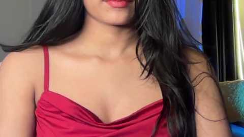 Media: Video of a young woman with long black hair, wearing a red spaghetti-strap dress, smiling slightly. Background shows a dimly lit room with a window and a brown sofa.