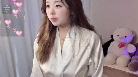 Video of a young Asian woman with long brown hair, wearing a white robe, standing in a softly lit room with a teddy bear and a heart-covered curtain in the background.