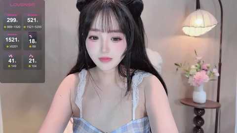 Video of a young East Asian woman with pale skin, long black hair styled in buns, wearing a light blue plaid dress, sitting in a softly lit room with a floral arrangement and a standing lamp.