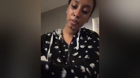 Media: A video of a Black woman with medium brown skin, wearing a black hoodie with white bear patterns, looking down. The background is dimly lit with blurred objects.