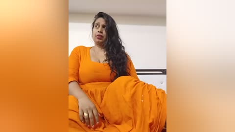 Video of a South Asian woman with long, dark hair, wearing an orange saree, seated in a modern, minimalist room with white walls.