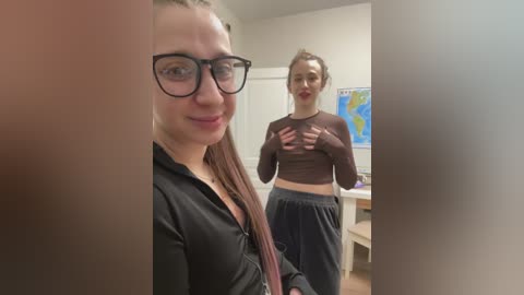 Media: Video of two young women in a room: one with glasses and long brown hair, wearing a black blouse, the other with blonde hair in a ponytail, wearing a brown long-sleeve top and loose pants.