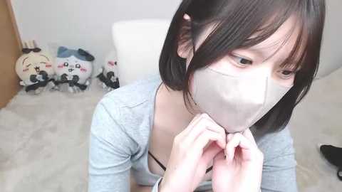 Media: A video of an Asian woman with short black hair, wearing a light gray cardigan, and a white mask, kneeling on a light-colored rug. In the background, stuffed panda toys are visible.