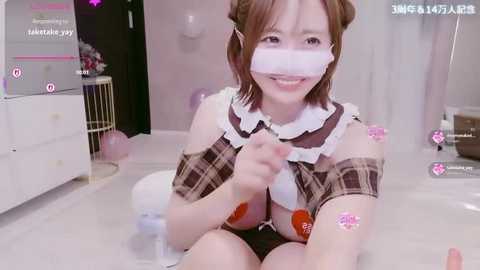 Media: Video of an Asian woman with light skin, short brown hair, wearing a plaid maid outfit, face mask, and heart stickers, sitting on a white floor, in a room with pink walls and a white dresser.
