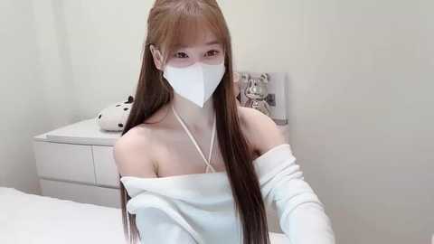 A video of an East Asian woman with long brown hair, wearing a white off-shoulder dress, face mask, and gloves, sitting on a white bed in a minimalist room.