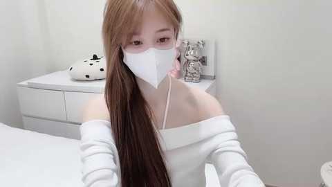 Video of an Asian woman with long brown hair, wearing a white off-shoulder top, face mask, and earphones, sitting on a white bed in a minimalist bedroom with white furniture.