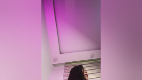 A video of a woman with long, dark hair standing in front of a purple-lit room with a white ceiling, a vent, and a window with horizontal blinds.