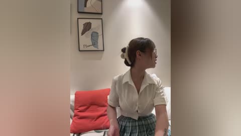 Video of a young woman with fair skin, brown hair in a bun, wearing a white blouse and green plaid skirt, seated on a bed with red and green pillows, against a beige wall with framed abstract art.